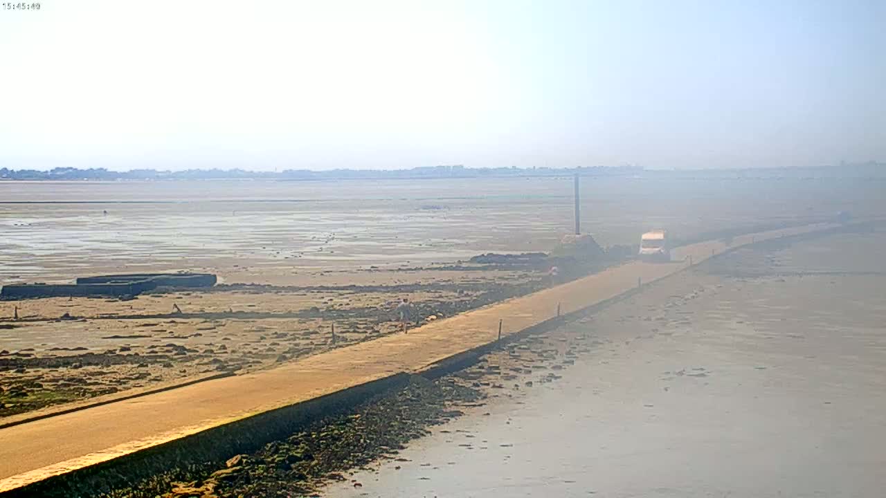 A long, light-brown road stretches across a hazy, flat expanse of mudflats toward a distant shoreline under a bright, hazy sky.