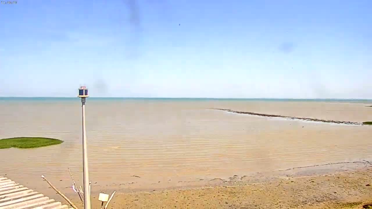 A mostly sunny day shows a calm, shallow body of water with a sandy shore and a tall pole with a light on top.