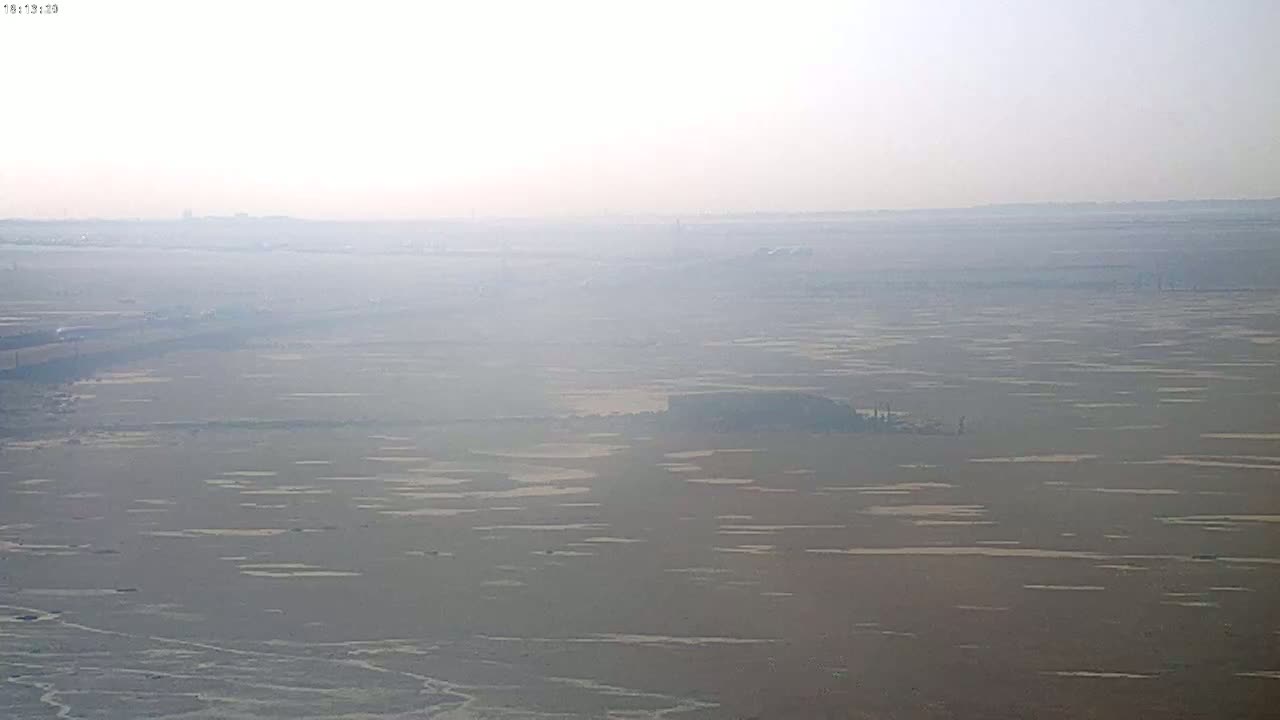 A hazy, distant view of a flat, mostly brown landscape with patches of lighter color, possibly water, under a light, overcast sky.