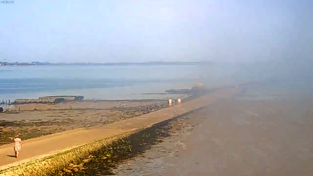 A hazy, sunny day reveals several people walking along a path bordering a calm body of water and a wide expanse of sandy beach.