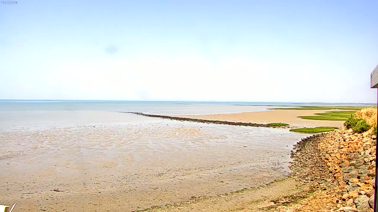 A rocky shoreline meets a calm, shallow, light-brown sea under a clear, bright sky.