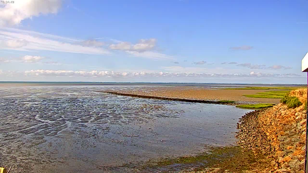 A wide expanse of tidal flats and shallow water stretches to a calm sea under a partly cloudy blue sky.