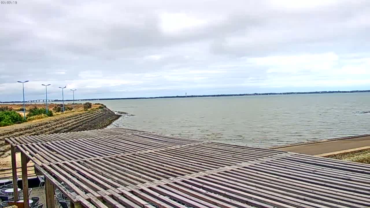 A wooden pergola overlooks a calm body of water under a cloudy sky, with a distant bridge visible on the horizon.