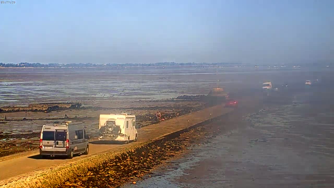 Several vehicles, including campervans, travel along a narrow road that extends across a wide expanse of exposed mudflats under a clear, sunny sky.