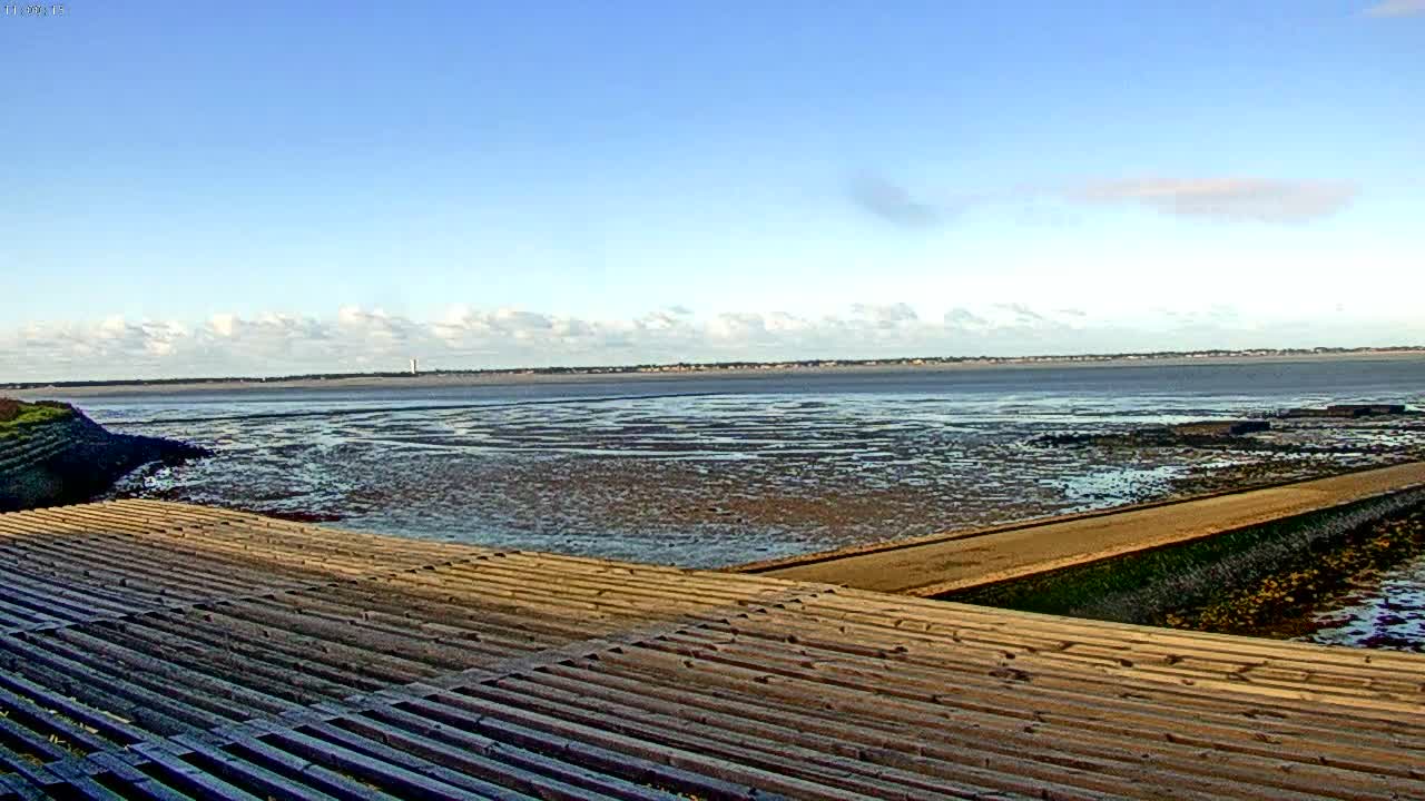 An expansive tidal bay, revealing intricate patterns of wet mudflats at low tide, stretches under a partly cloudy blue sky, with a wooden-slatted structure and concrete path in the foreground leading towards a distant shoreline.