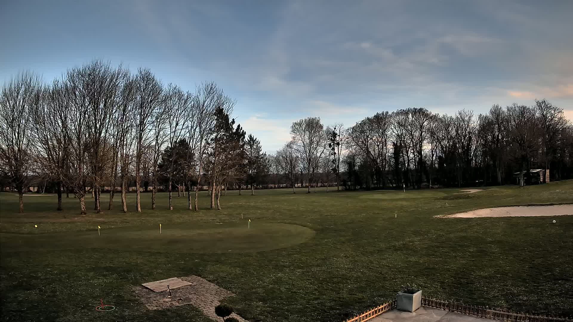 A mostly barren, green golf course with leafless trees under a partly cloudy sky.
