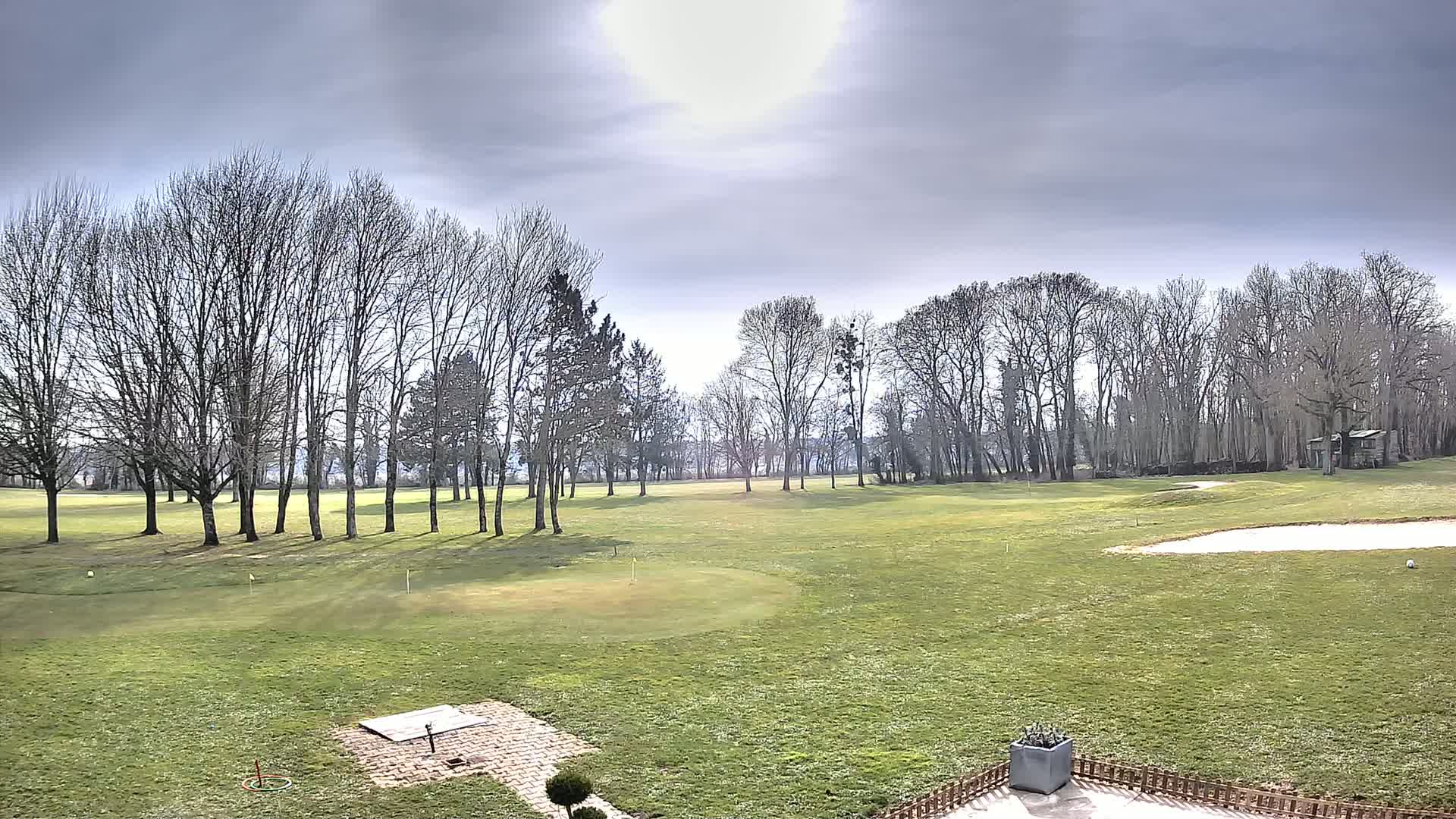A mostly sunny day with overcast skies shows a view of a grassy golf course with bare trees lining the edge of the fairway.