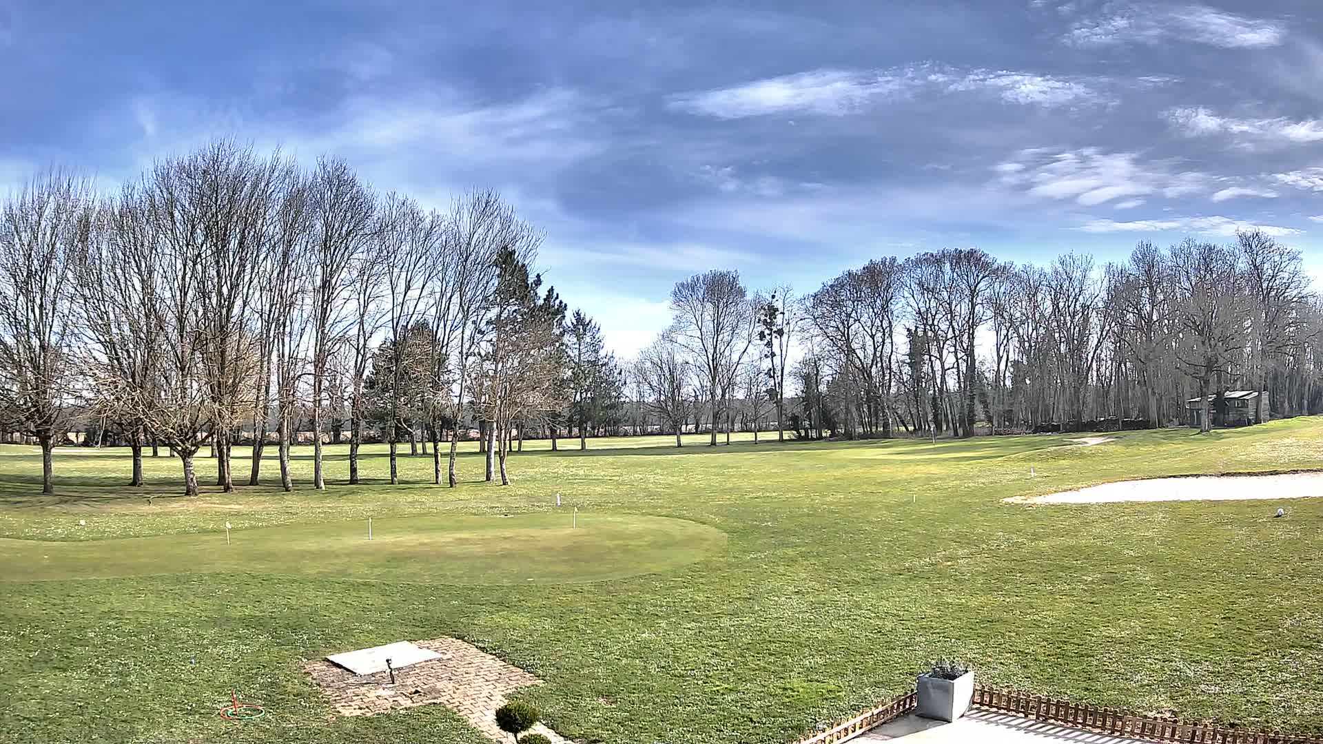 A mostly sunny day reveals a sprawling green golf course dotted with bare trees and a sand trap, all under a bright blue sky with scattered clouds.