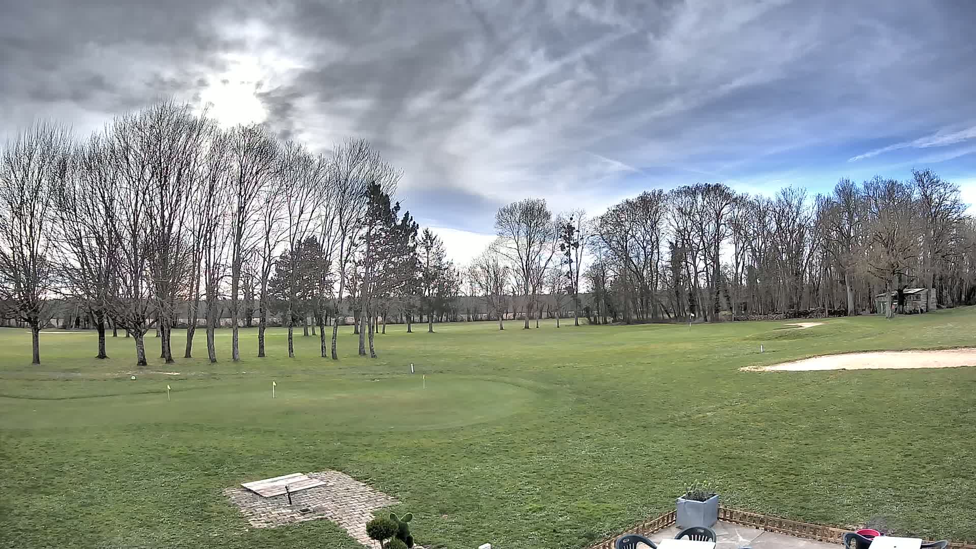 A partly sunny day with cloudy skies overlooks a golf course with leafless trees lining the perimeter and a sand trap visible in the distance.