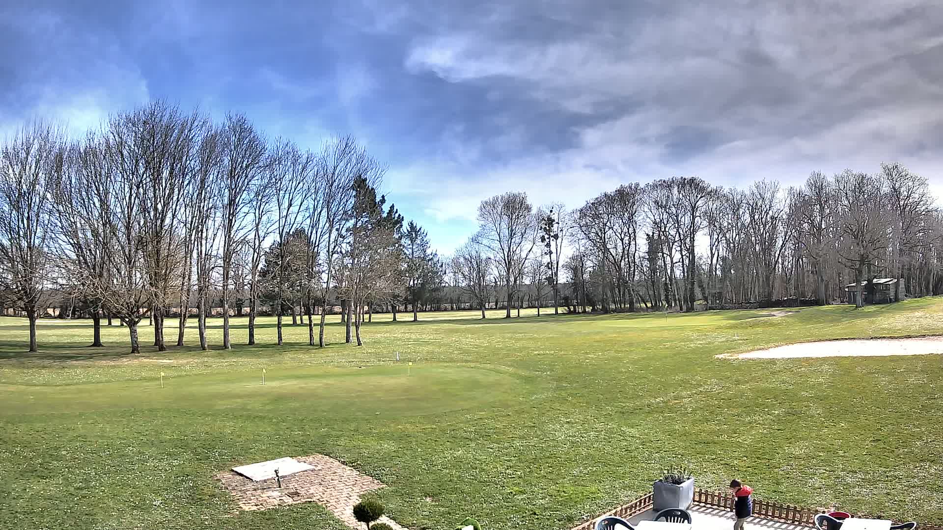 A mostly sunny day with partly cloudy skies overlooks a sprawling green golf course dotted with bare trees and a sand trap.