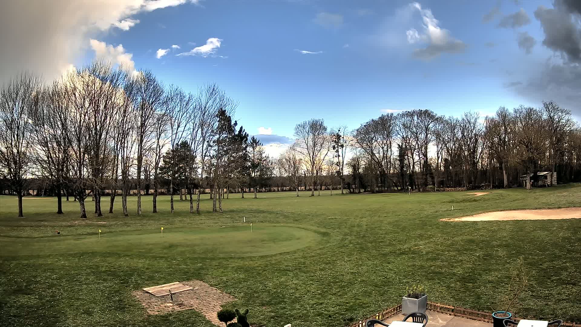 A partly cloudy sky overlooks a short, grassy golf course with leafless trees and a sand trap.