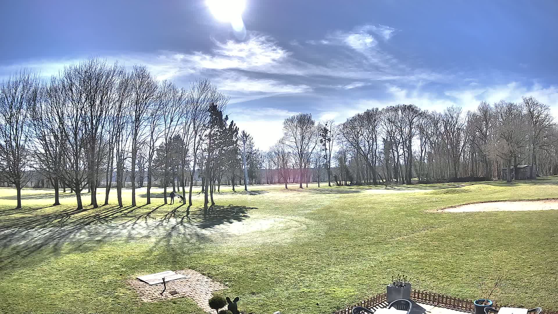 A sunny day reveals a golf course with leafless trees casting shadows on a grassy fairway, a sand trap, and a small structure in the distance.