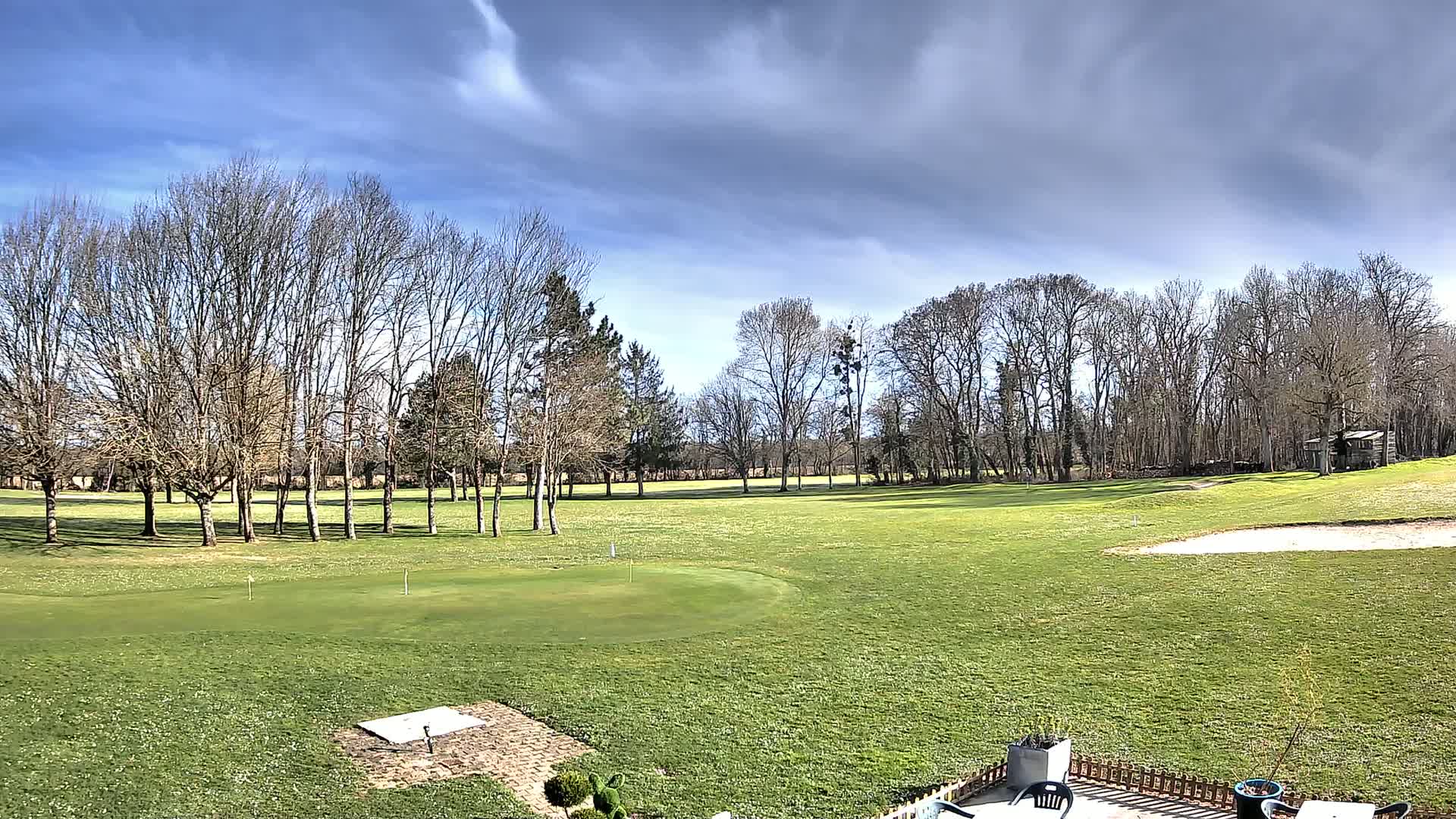 A mostly sunny day with partly cloudy skies overlooks a putting green and fairway on a golf course surrounded by bare deciduous trees.
