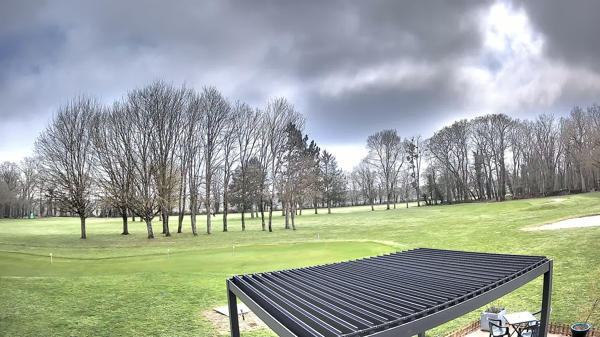 A green golf course with bare trees under a cloudy sky is partially visible from beneath a dark pergola.