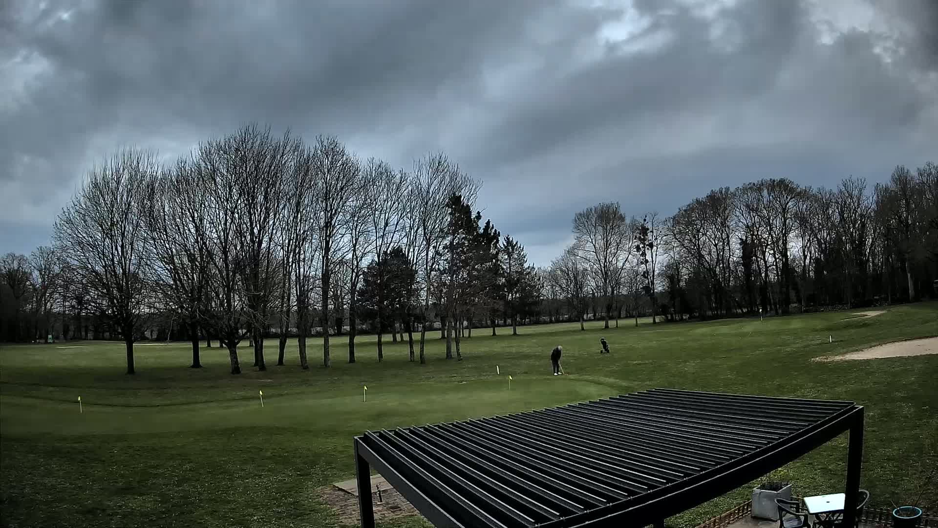Two golfers are on a green putting area under a cloudy sky, with bare trees in the background and a dark slatted pergola in the foreground.