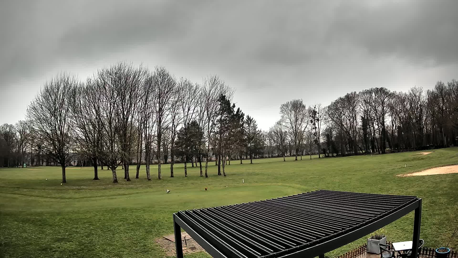 A green golf course with bare trees under a cloudy sky is partially visible from beneath a slatted pergola.