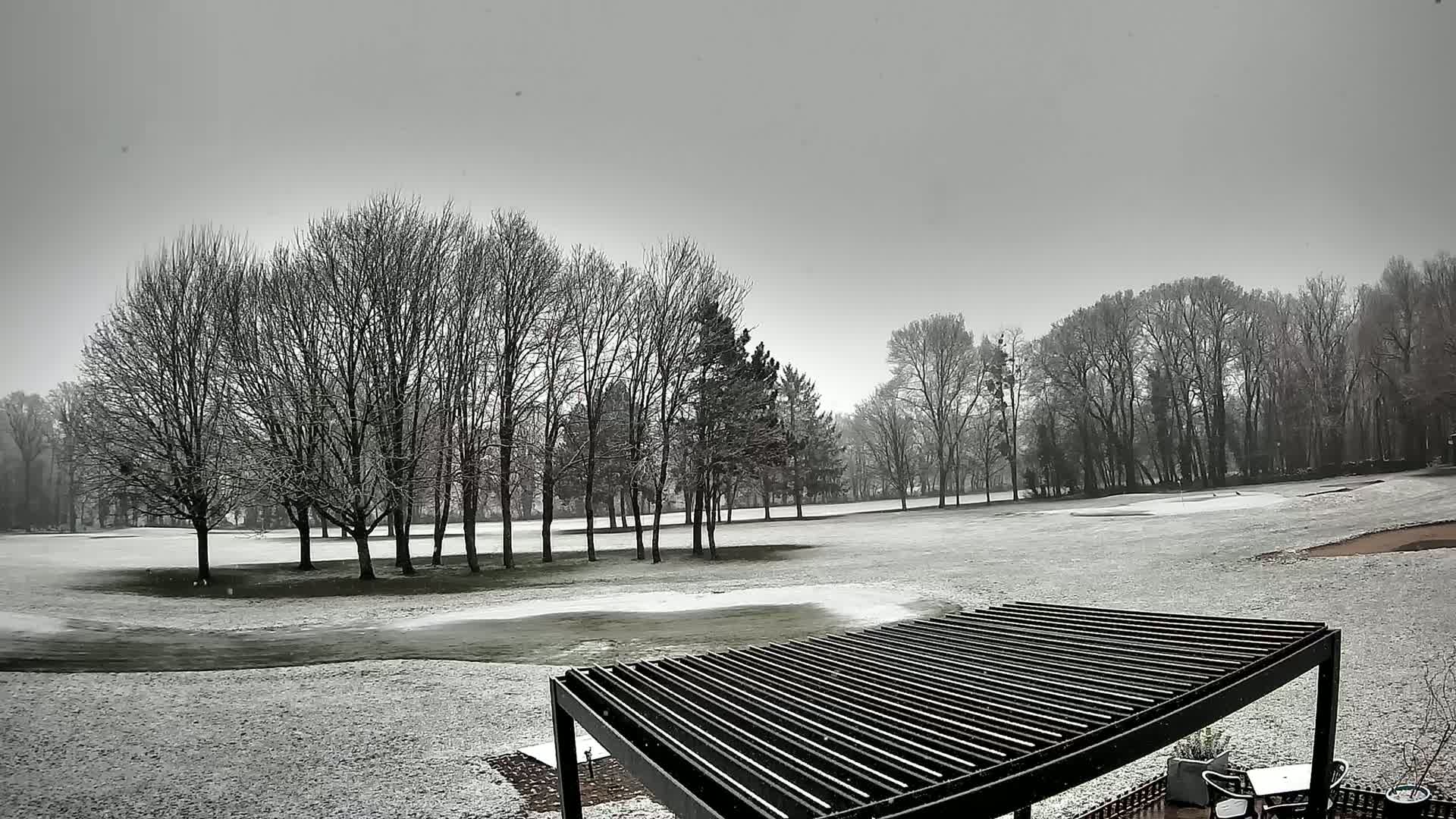 A snow-covered golf course with leafless trees under a gray sky is viewed from behind a slatted pergola.
