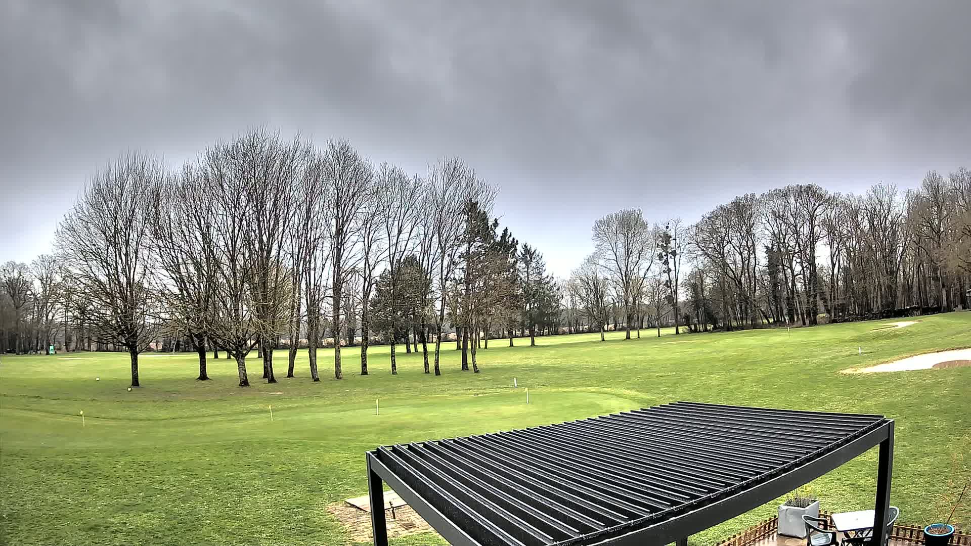 A sprawling green golf course with bare trees under a cloudy sky is partially obscured by a modern black pergola.