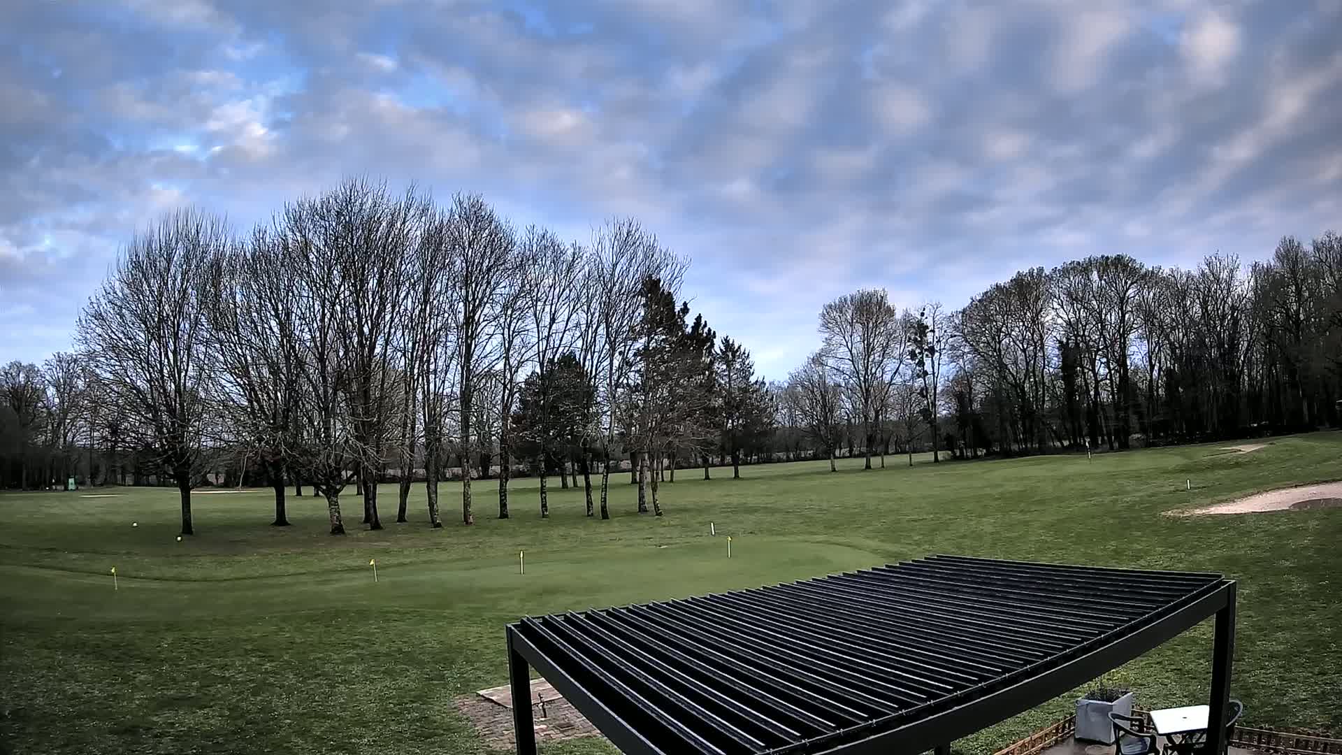A mostly cloudy sky overlooks a green golf course with bare trees and a black pergola.