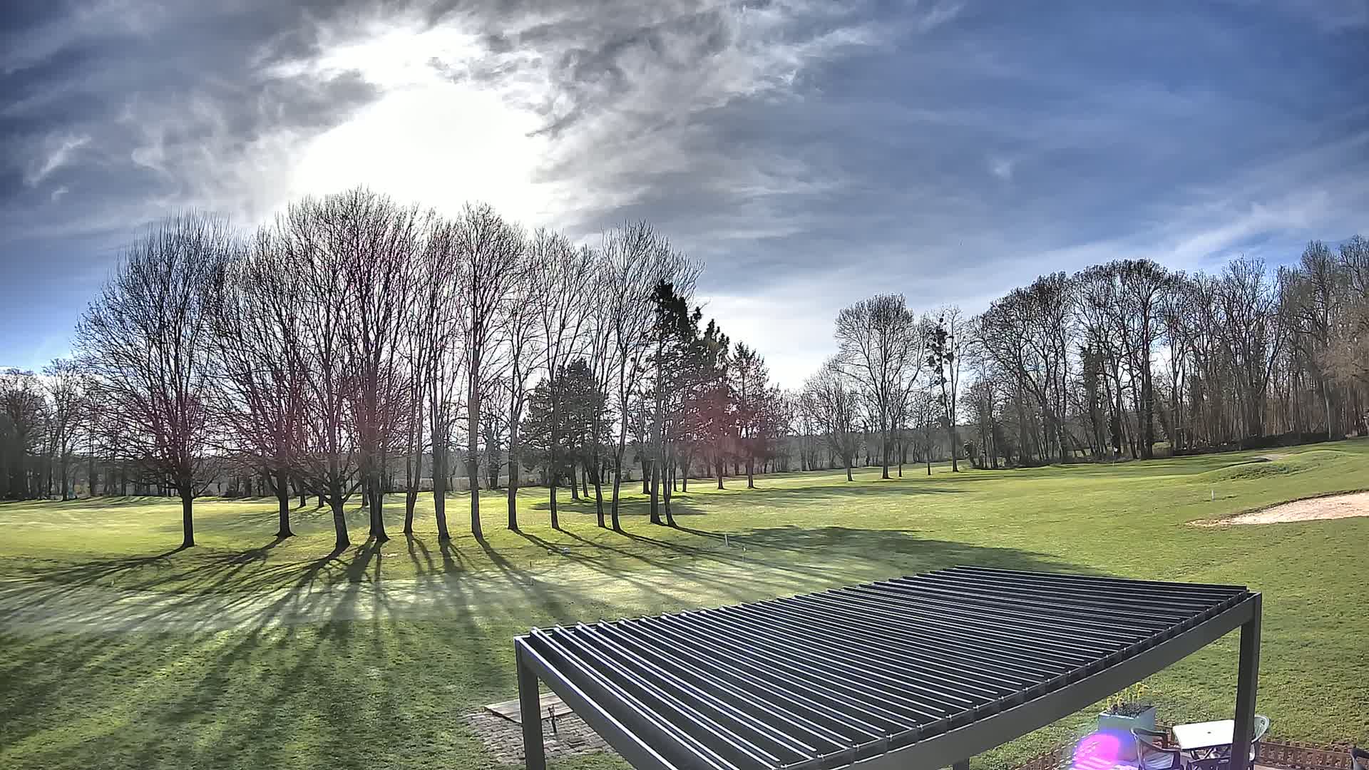 A mostly sunny day reveals a golf course with leafless trees casting long shadows on a green lawn, partially obscured by a dark slatted pergola in the foreground.