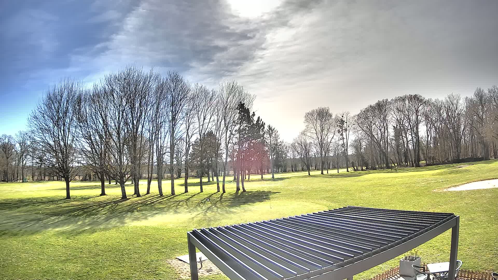 A mostly sunny day with partly cloudy skies over a green golf course dotted with bare deciduous trees and a modern pergola in the foreground.