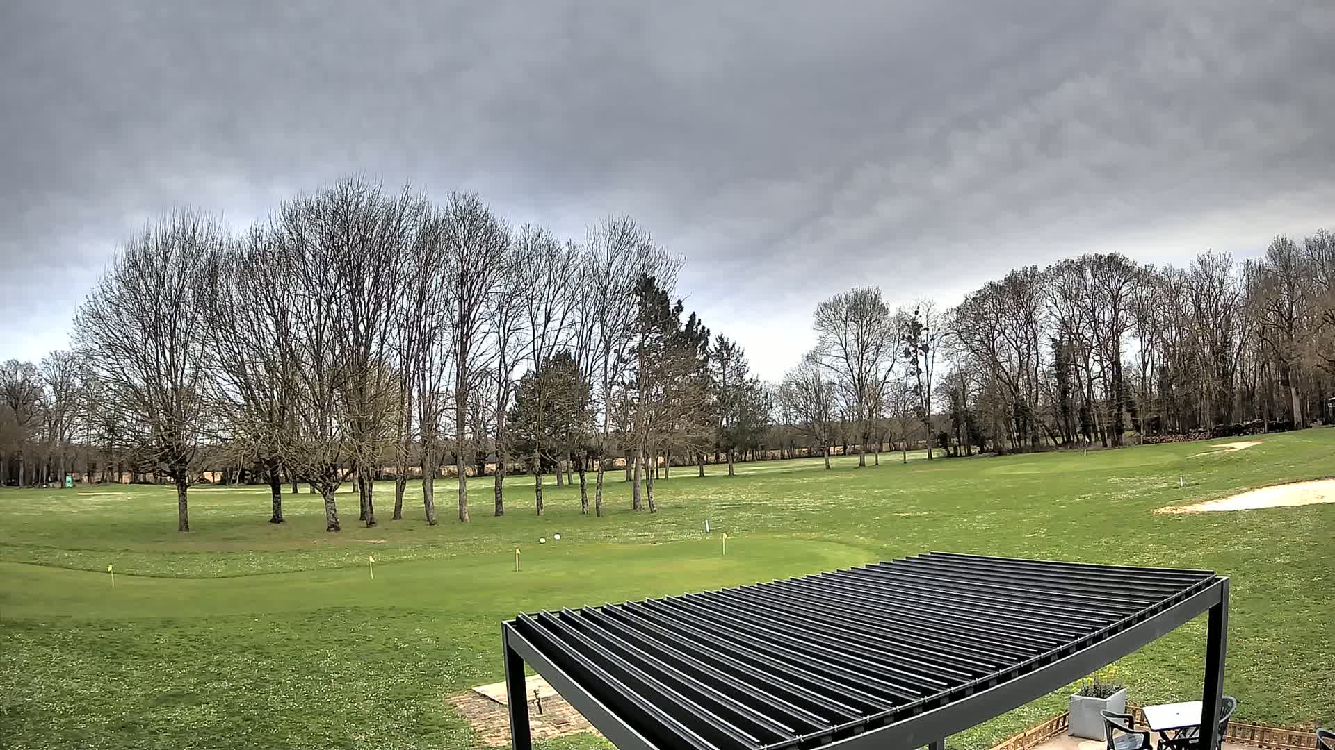 A golf course with bare trees under a cloudy sky is partially visible from beneath a dark pergola.