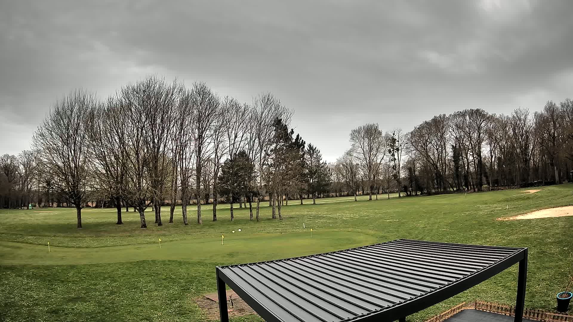 A golf course with bare trees under a cloudy sky is partially visible beneath a dark gray pergola.