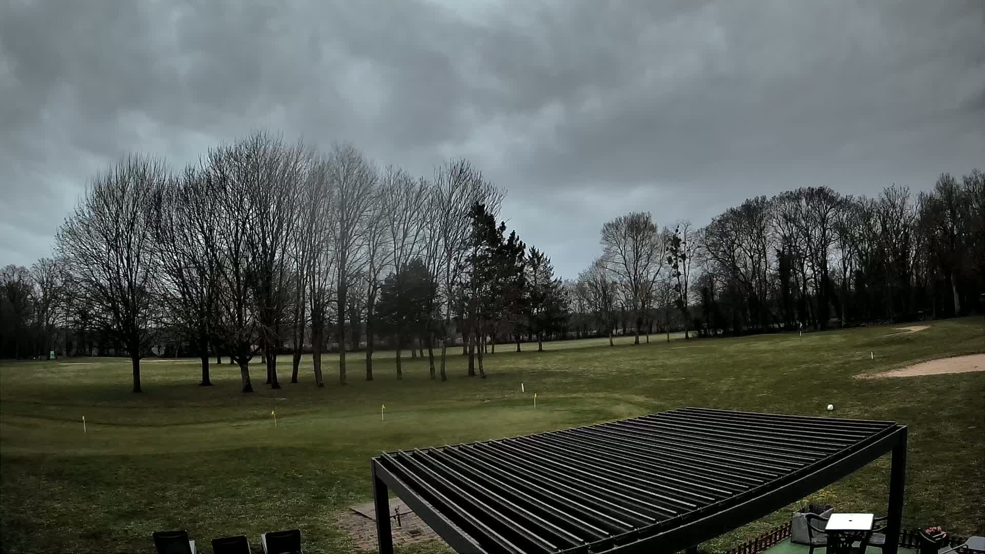 A green golf course with bare trees under a cloudy sky is partially visible from under a slatted pergola.