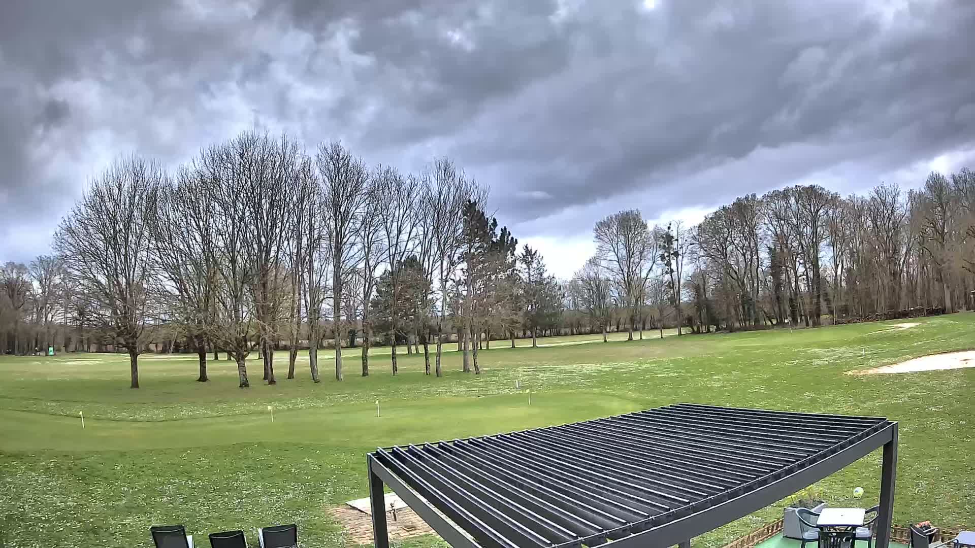 A view of a golf course with bare trees under a cloudy sky shows a grassy putting green in the foreground and a sand trap visible in the background.