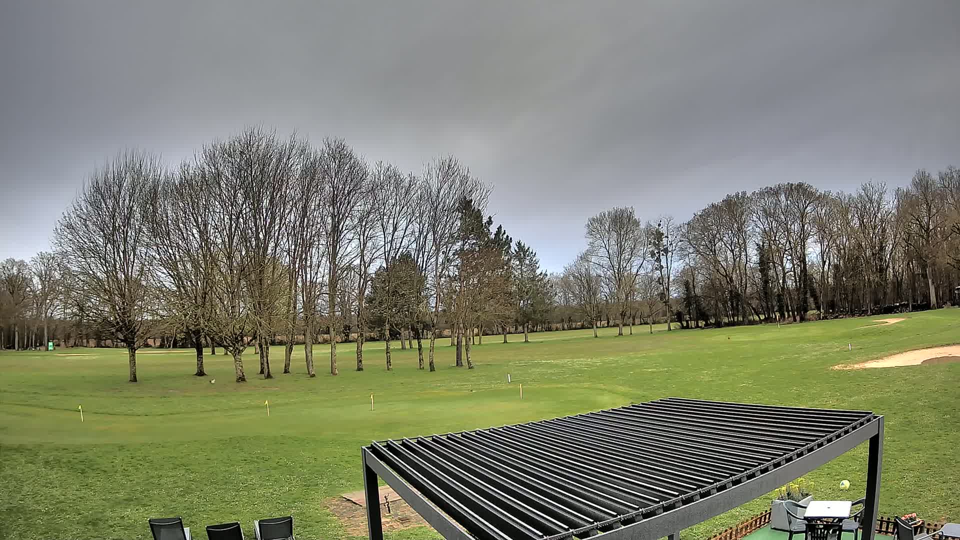 A green golf course with bare trees under an overcast sky, partially visible from under a dark metal pergola.