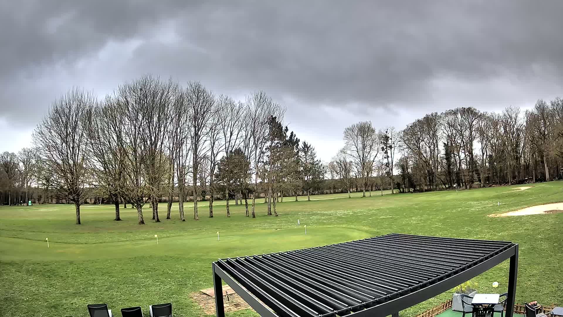 A view of a golf course under a cloudy sky, with bare trees lining the course and a dark pergola in the foreground.