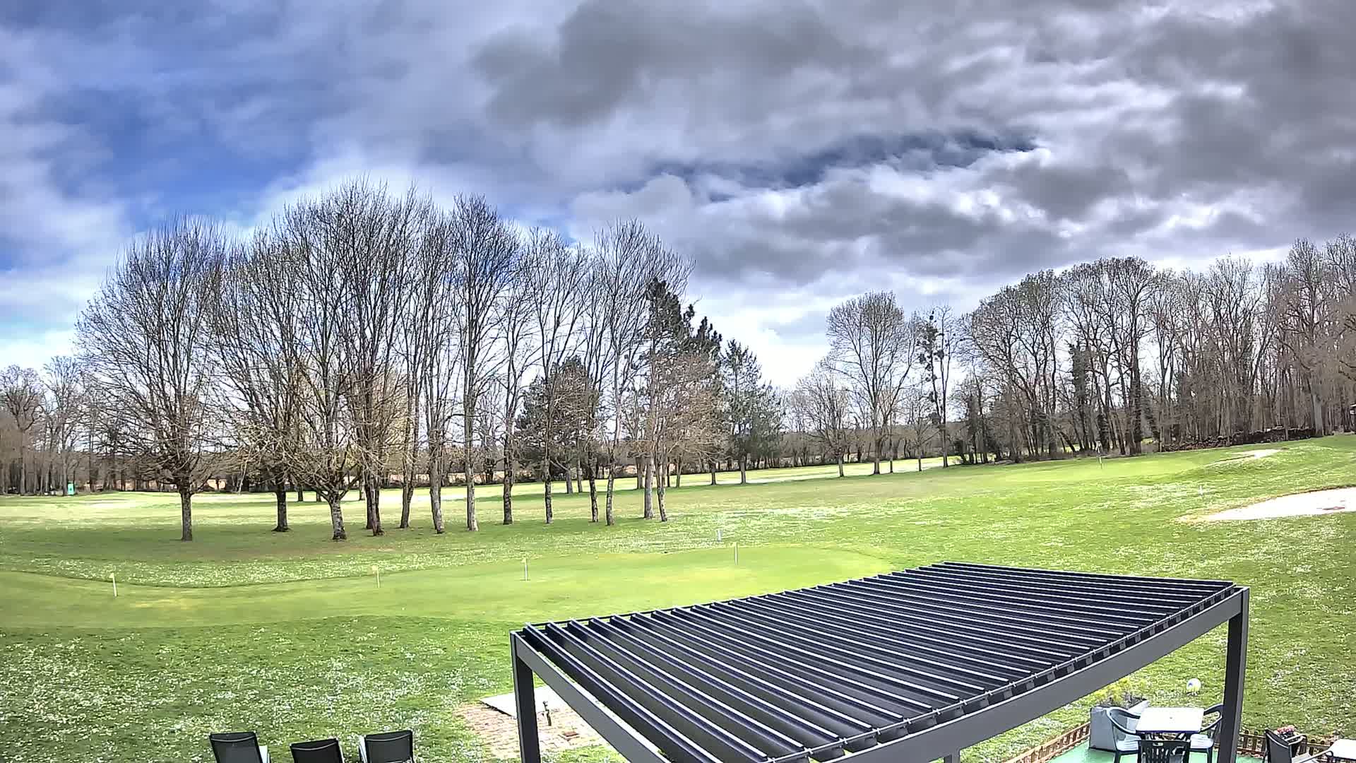 A mostly cloudy sky overlooks a green golf course dotted with bare trees and a dark-colored pergola.