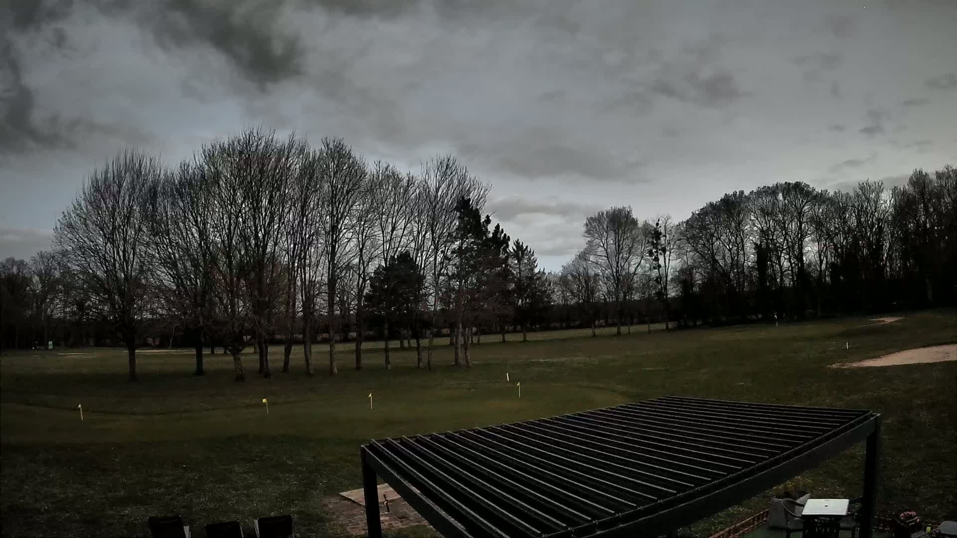A mostly barren, grassy field with leafless trees under a cloudy sky is viewed from above a slatted pergola.