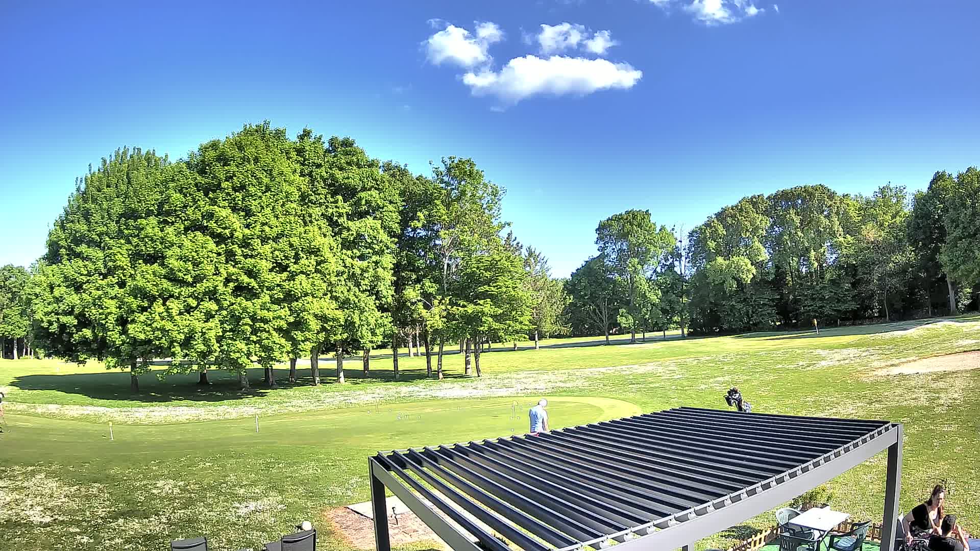 A sunny day on a golf course shows lush green grass, full green trees, a putting green, several people golfing, and a pergola.