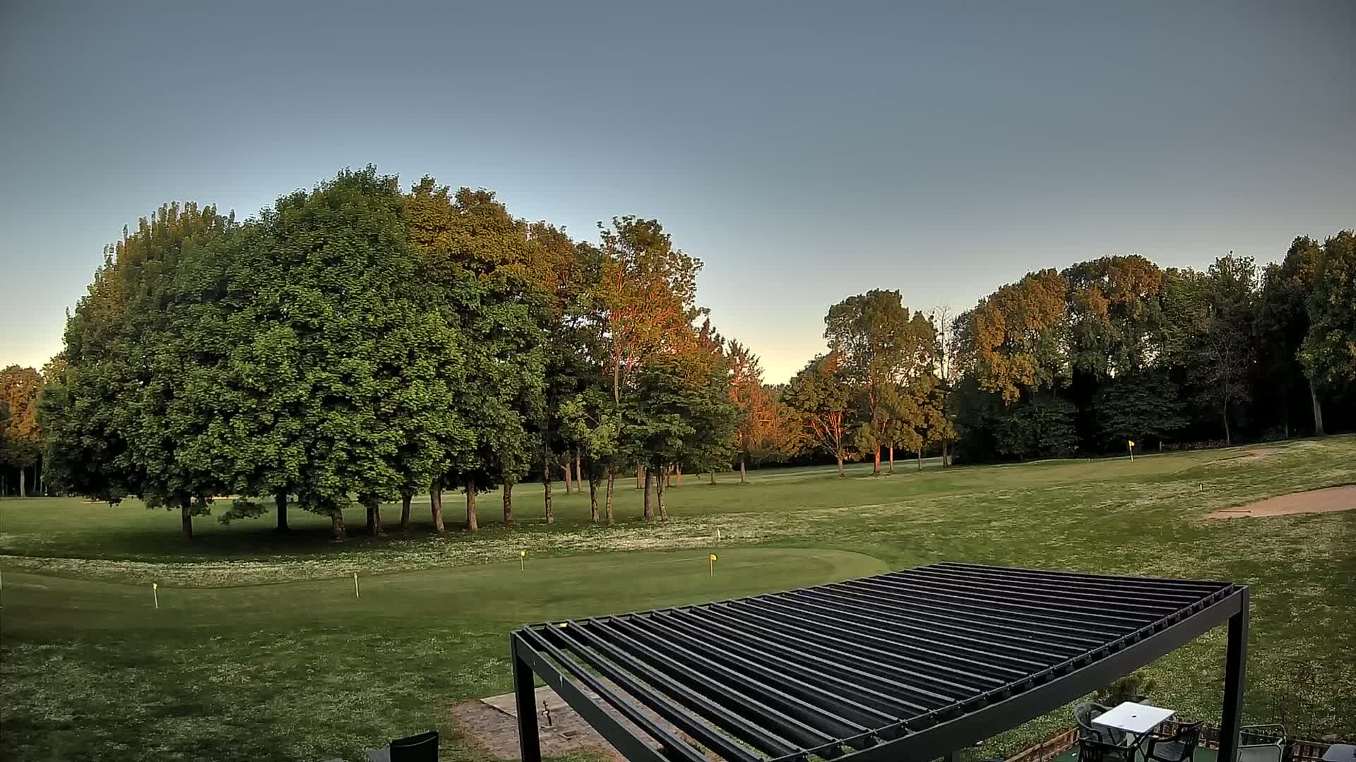 A golf course with green grass and several deciduous trees under a clear, light-blue sky at sunset is partially visible from beneath a black pergola.