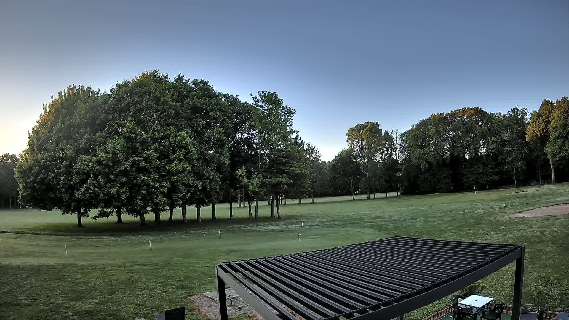 A grassy golf course with several leafy green trees under a clear blue sky is partially visible from behind a dark-colored pergola.
