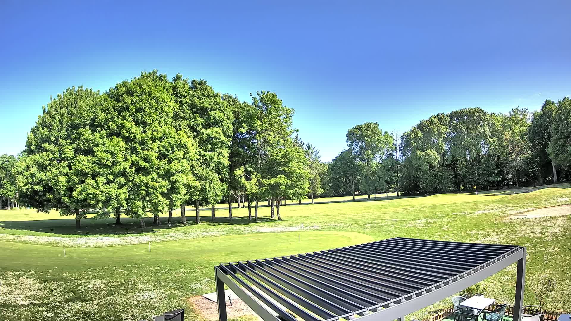 A grassy golf course with lush green trees under a clear blue sky is partially visible from beneath a dark metal pergola.