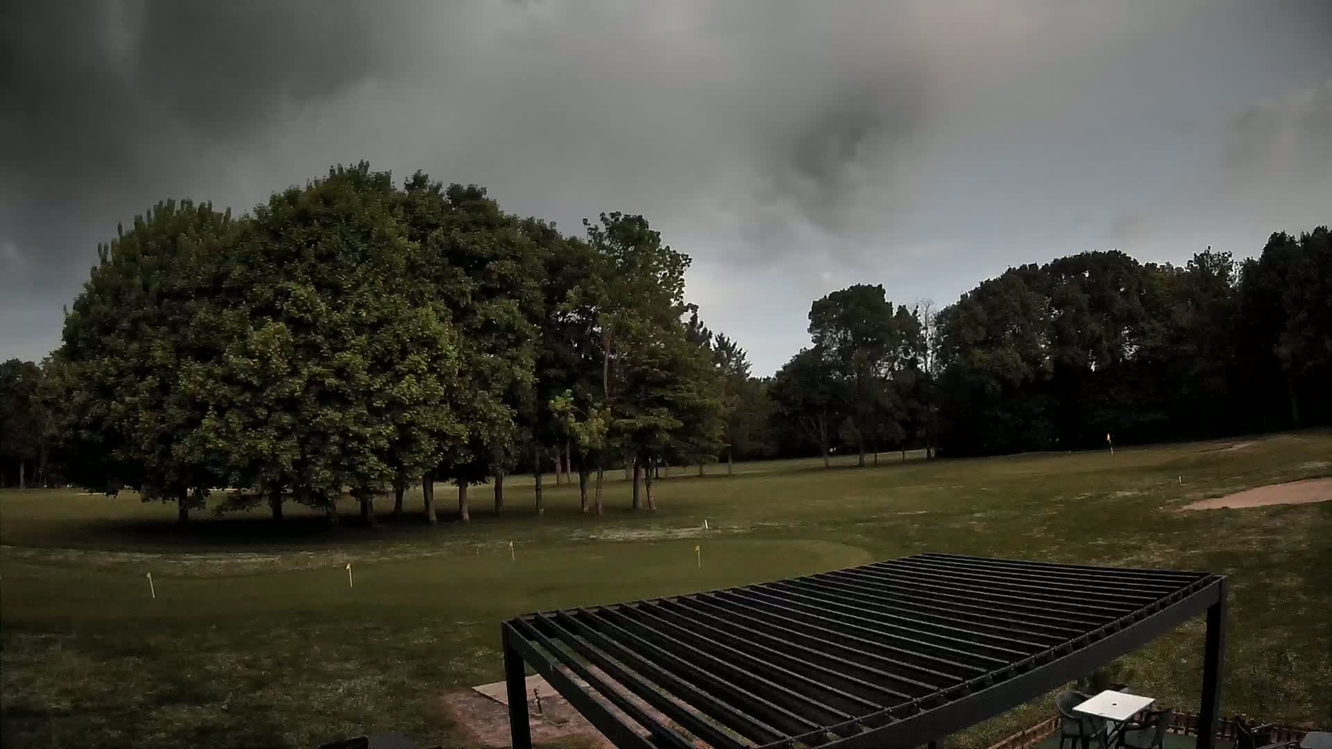 A putting green bordered by trees under a dark, cloudy sky is partially visible from above a black pergola.