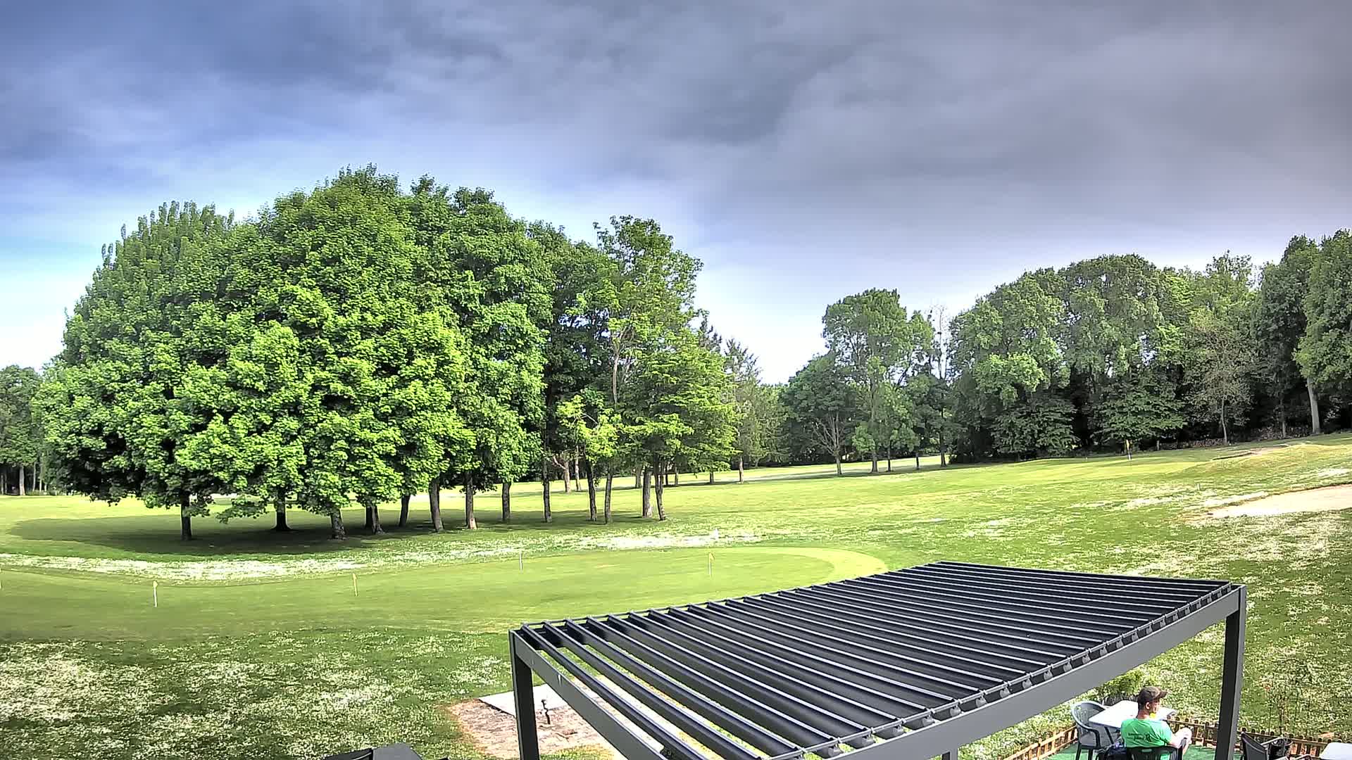 A person sits under a pergola overlooking a green golf course under a cloudy sky.