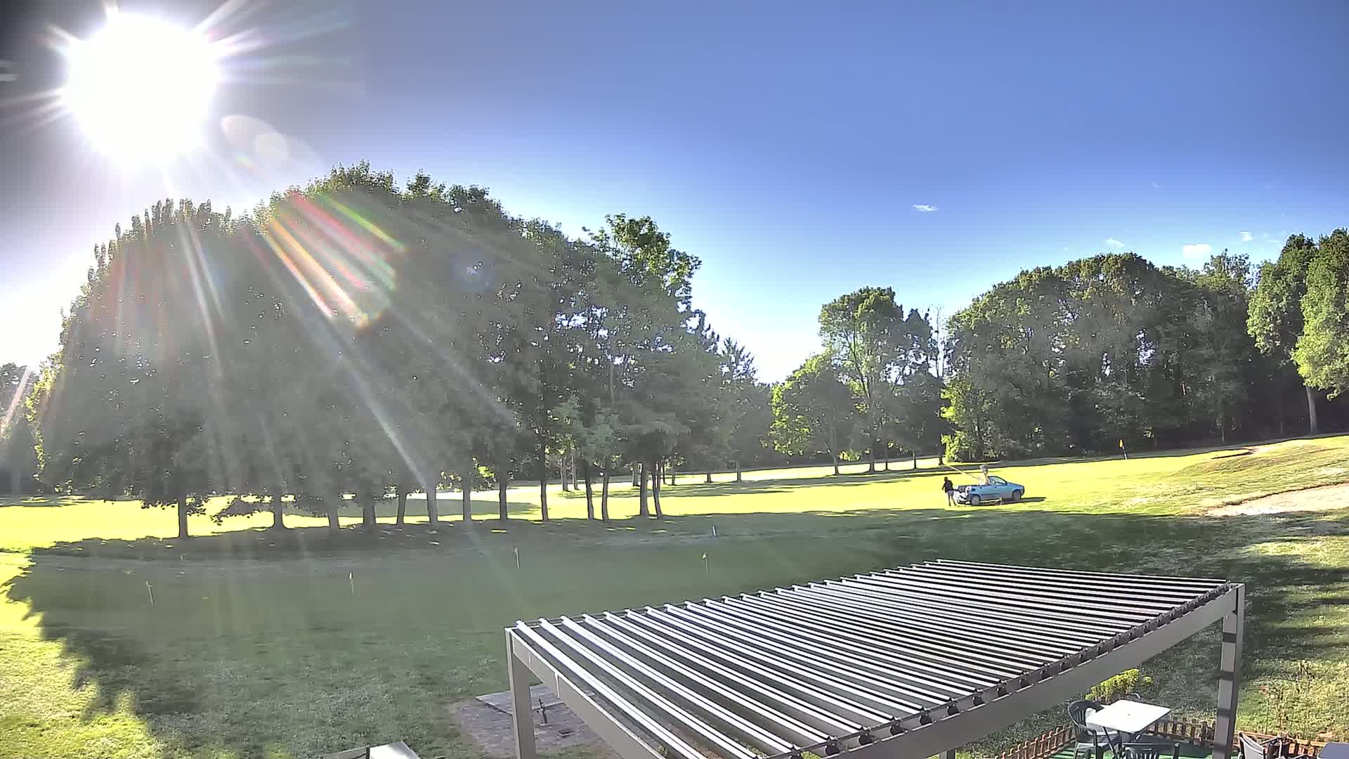 A sunny day on a golf course viewed from a patio with a pergola, showing a car and a person near the green.