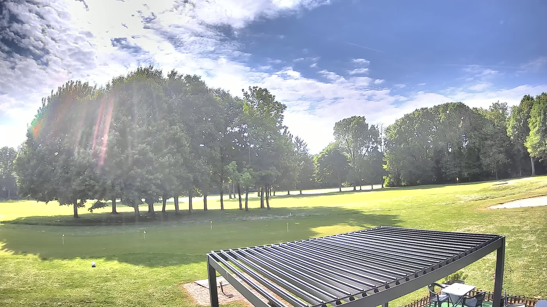 A partly sunny day on a golf course shows a putting green bordered by lush green trees, with a pergola in the foreground.