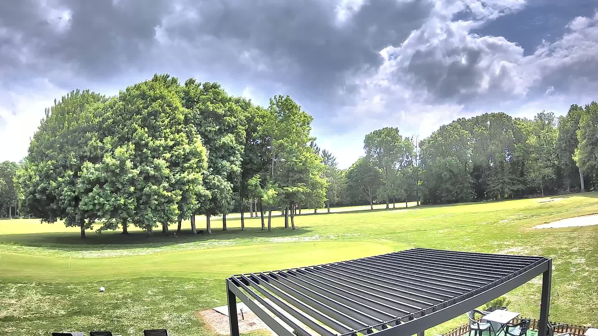 A partly cloudy sky overlooks a lush green golf course dotted with several large, full trees and a modern black pergola.