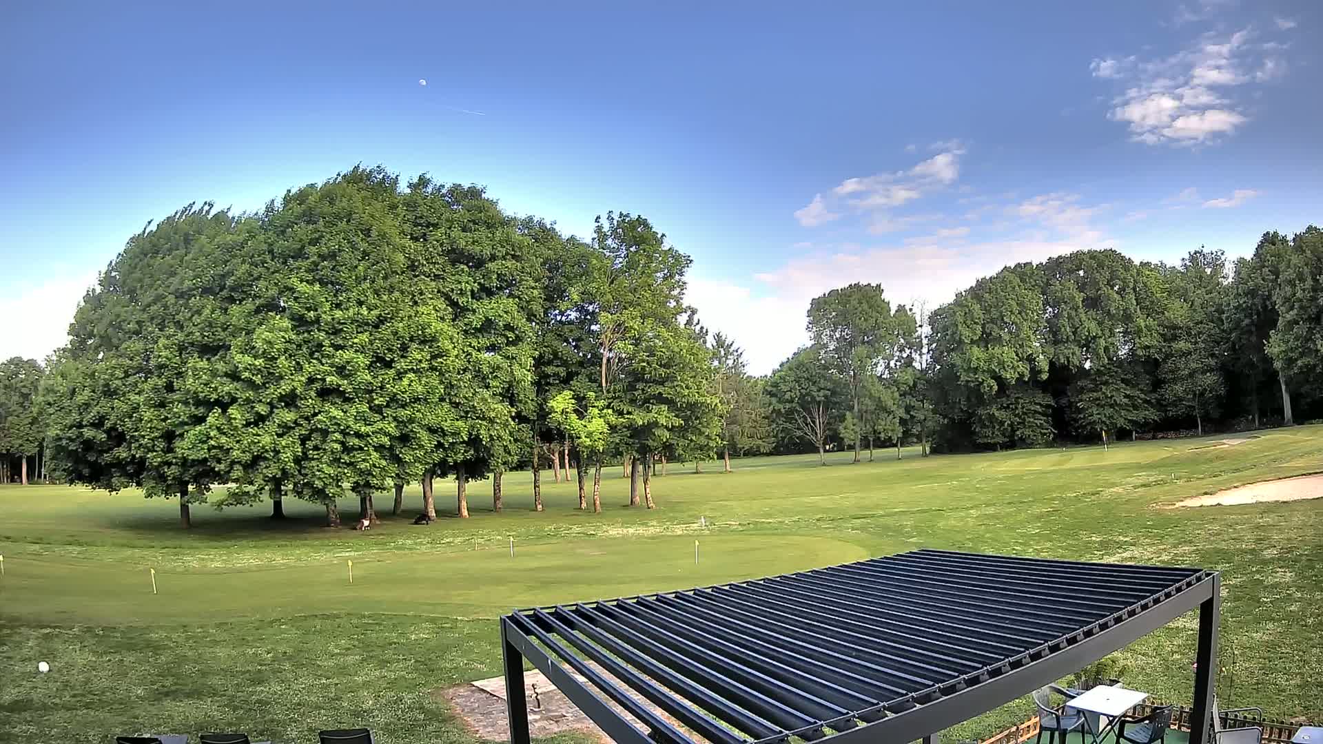 A partly cloudy blue sky overlooks a green golf course dotted with lush trees, and a dark pergola with a table and chairs sits in the foreground.