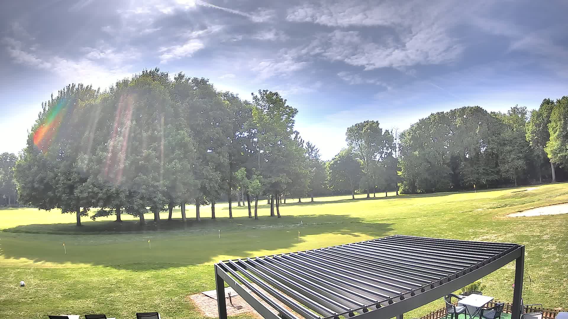 A sunny day reveals a verdant golf course bordered by lush green trees, partially obscured by a dark metal pergola.