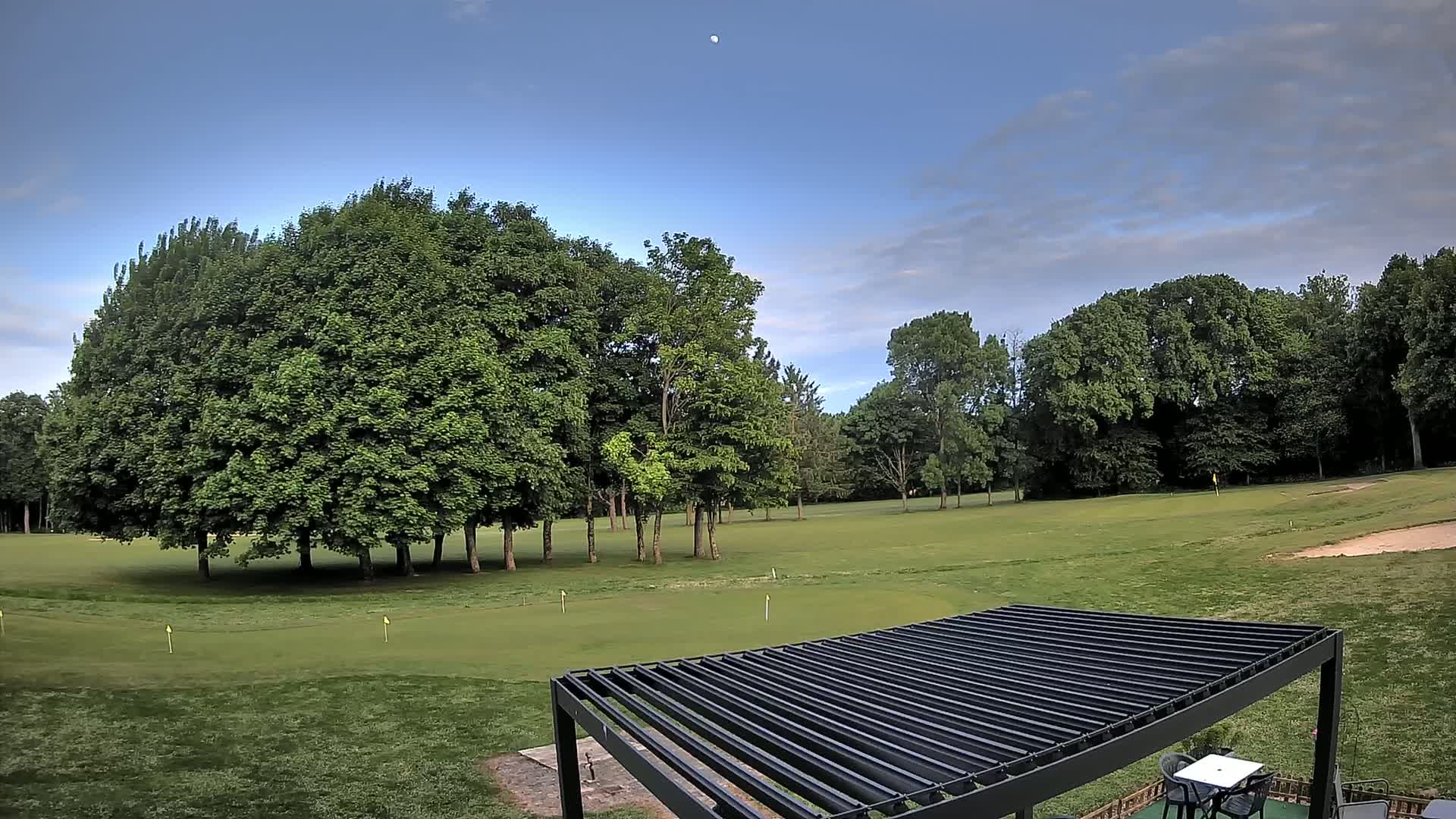 A mostly clear sky with a visible moon hangs over a green golf course bordered by lush, full trees, partially obscured by a dark-colored pergola.