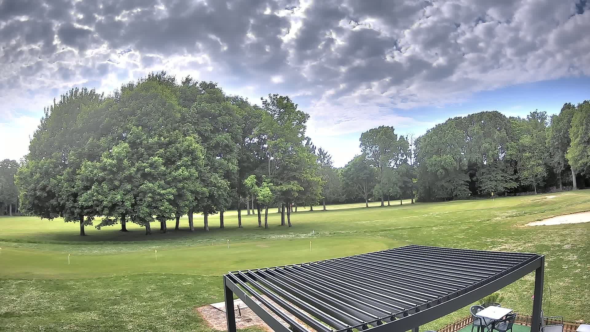 A partly cloudy sky overlooks a lush green golf course with several mature trees and a dark-colored pergola in the foreground.