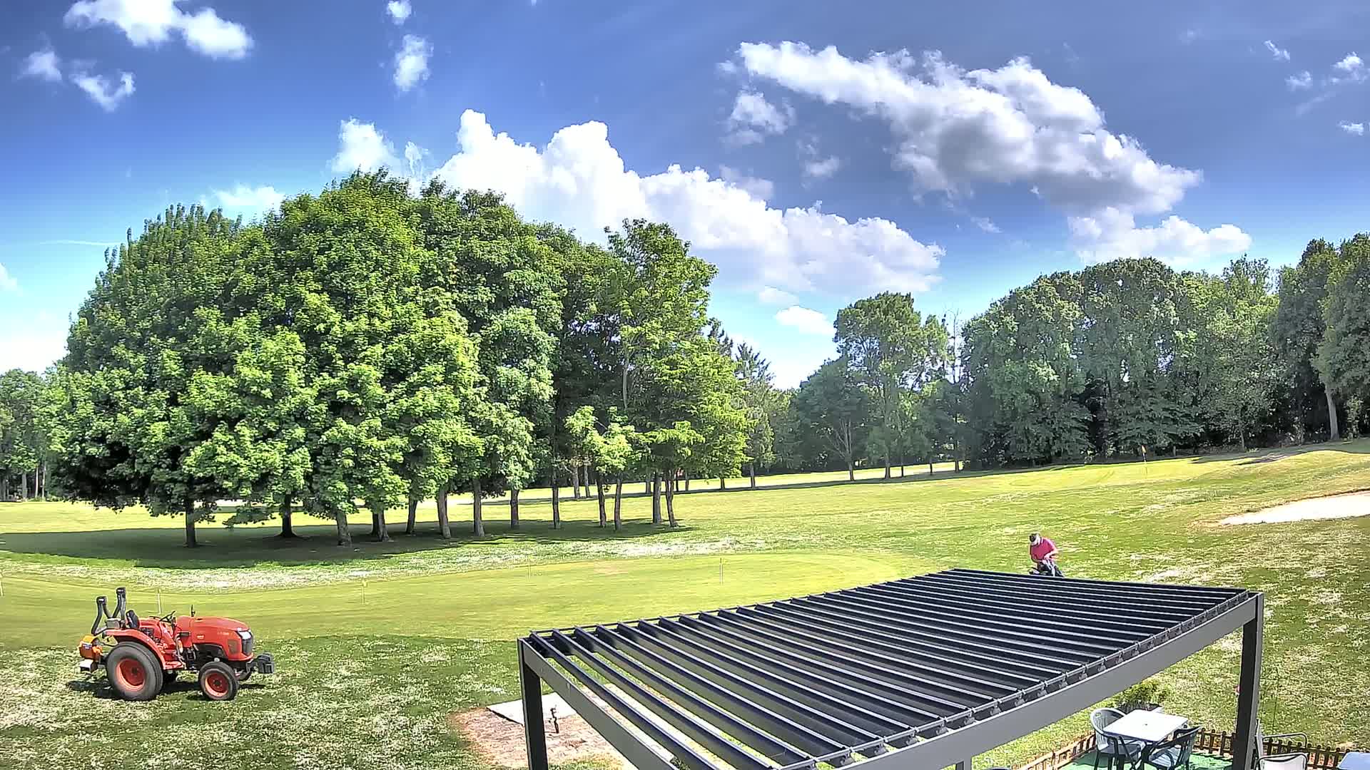 A sunny day with partly cloudy skies shows a golf course with a red tractor, a golfer, and a modern pergola.