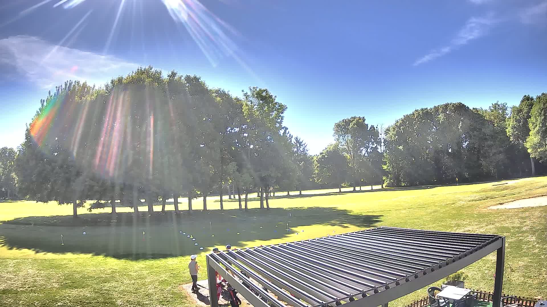 A sunny day on a golf course shows two people near a covered patio area overlooking a putting green surrounded by trees.