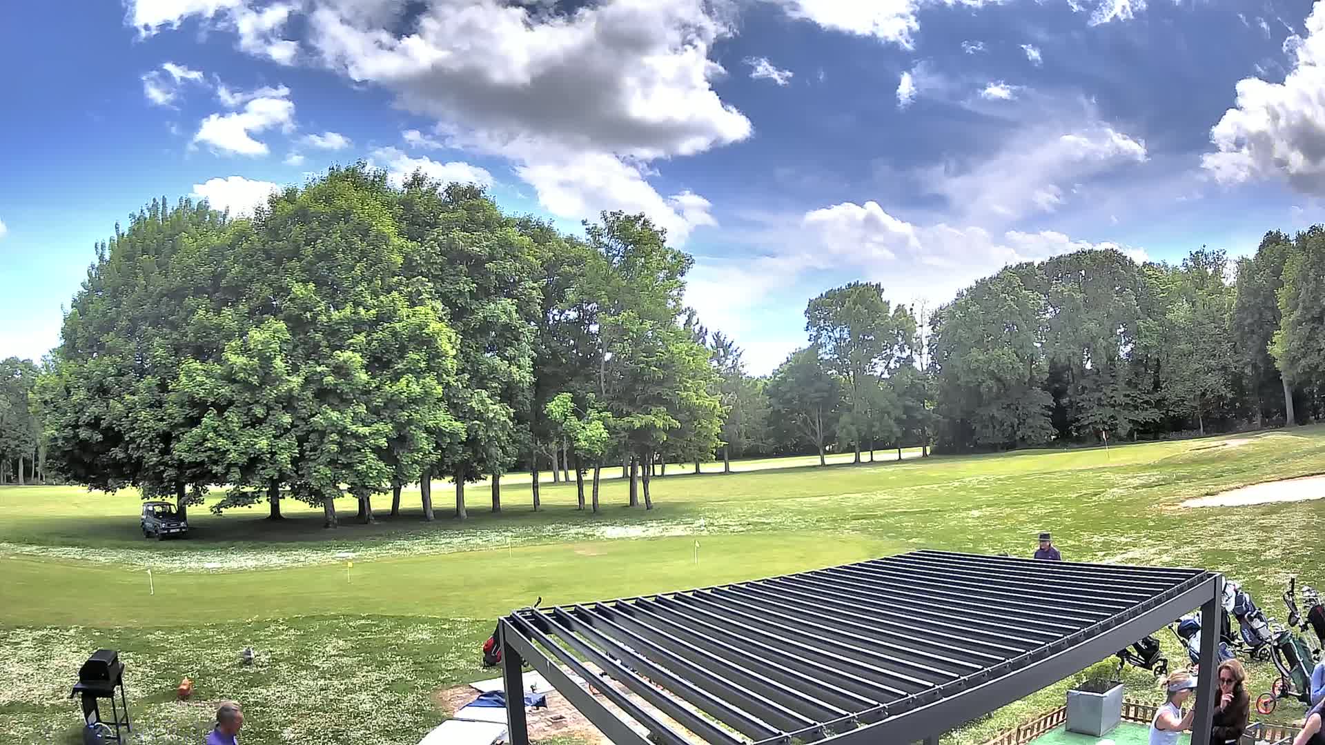 A partly cloudy sky overlooks a golf course with a few people near a partially constructed pergola.