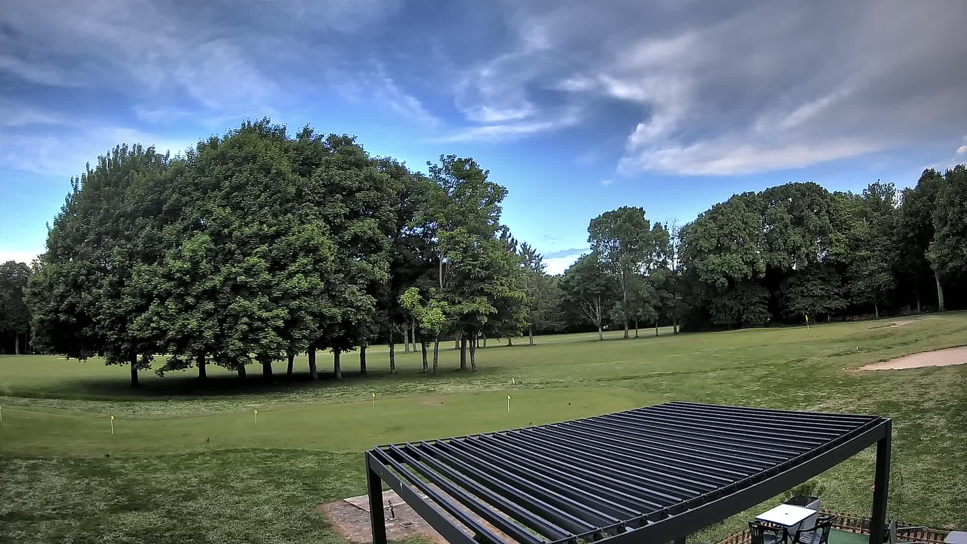 A mostly sunny sky with scattered clouds overlooks a green golf course dotted with lush trees and a modern black pergola.